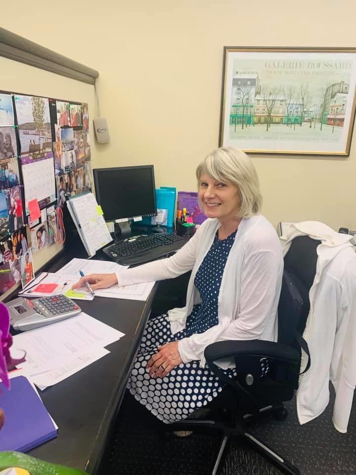 Woman at desk, smiling, in office setting. She wears a dress and cardigan. Computer and papers visible.