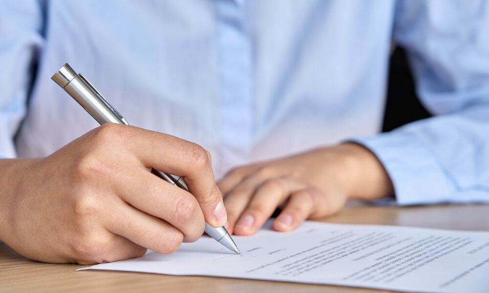 Person writing on paper with a pen; light blue shirt; wooden table.