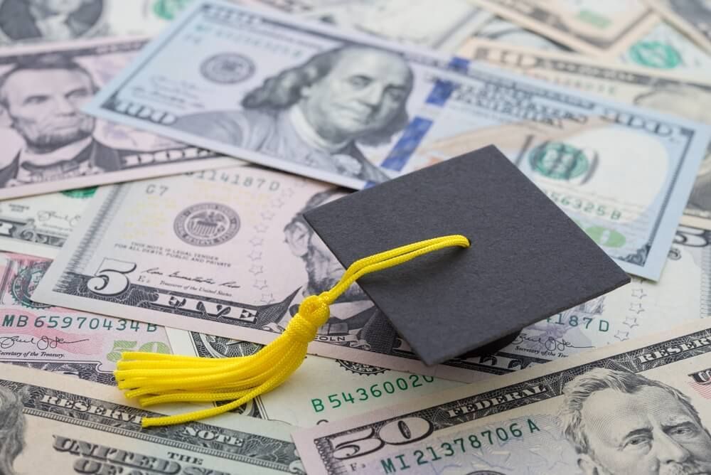 Graduation cap with yellow tassel on pile of US dollar bills.