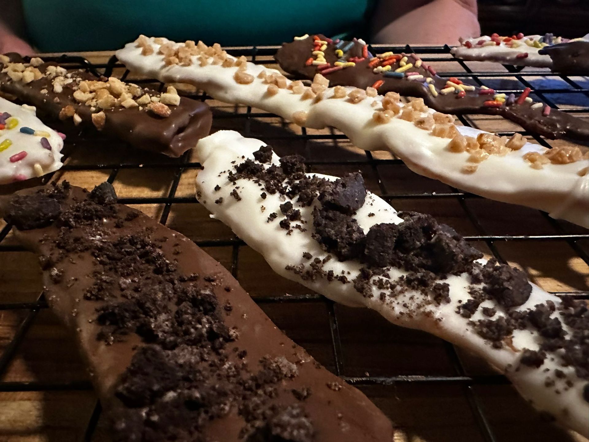 Chocolate-covered pretzel rods on a cooling rack; toppings include sprinkles, crushed Oreos, and peanuts.