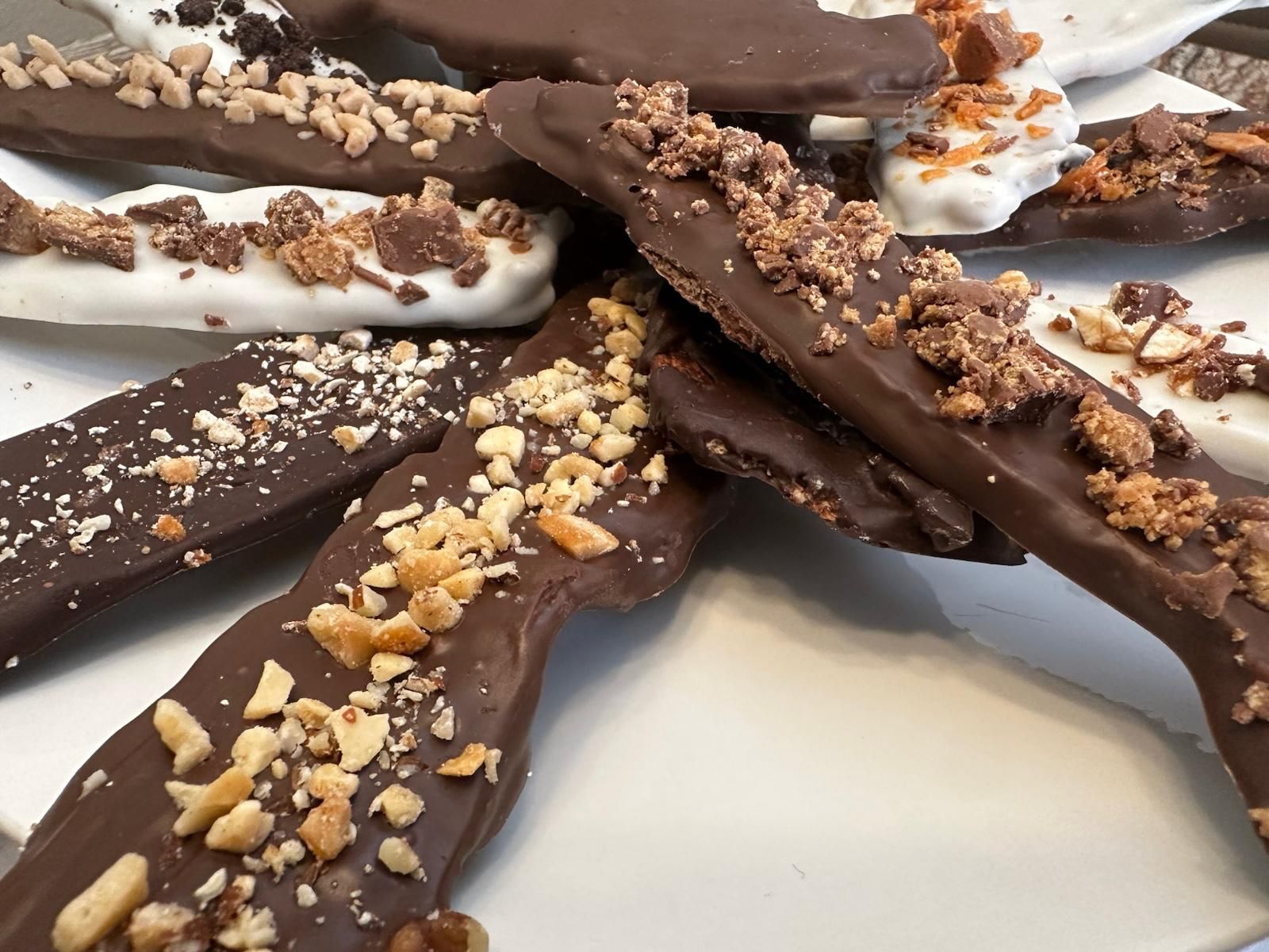 Chocolate-covered pretzel sticks, dark and white chocolate, topped with nuts and cookie crumbles on a white plate.