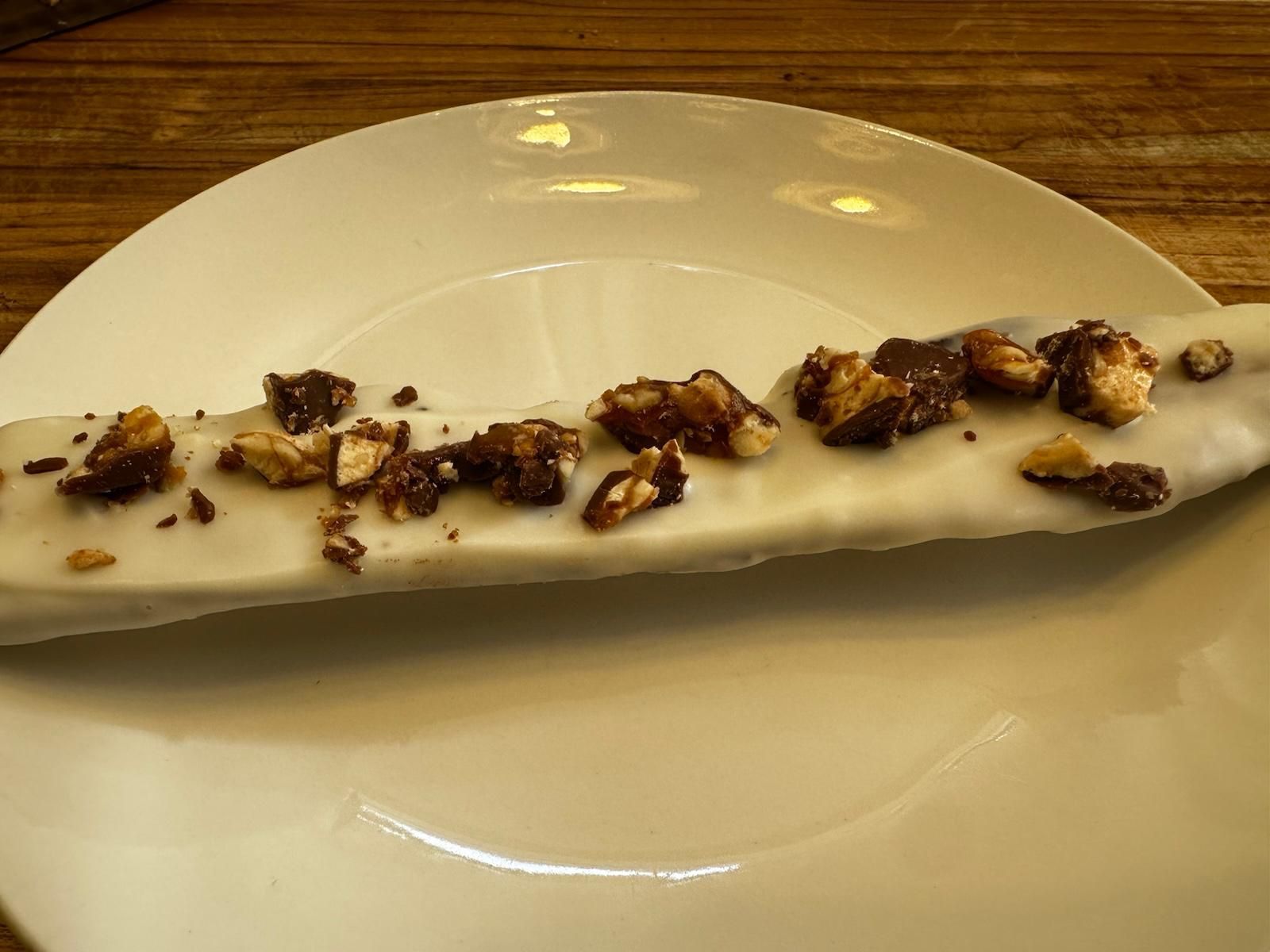 Banana dipped in white chocolate, topped with chocolate-covered nuts, on a white plate.