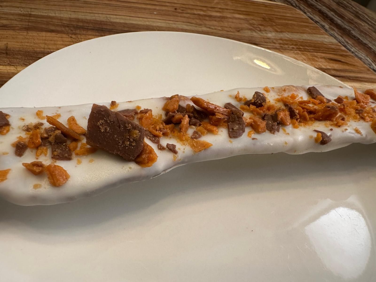 A long, white glazed pastry topped with chocolate chunks and honeycomb on a white plate.