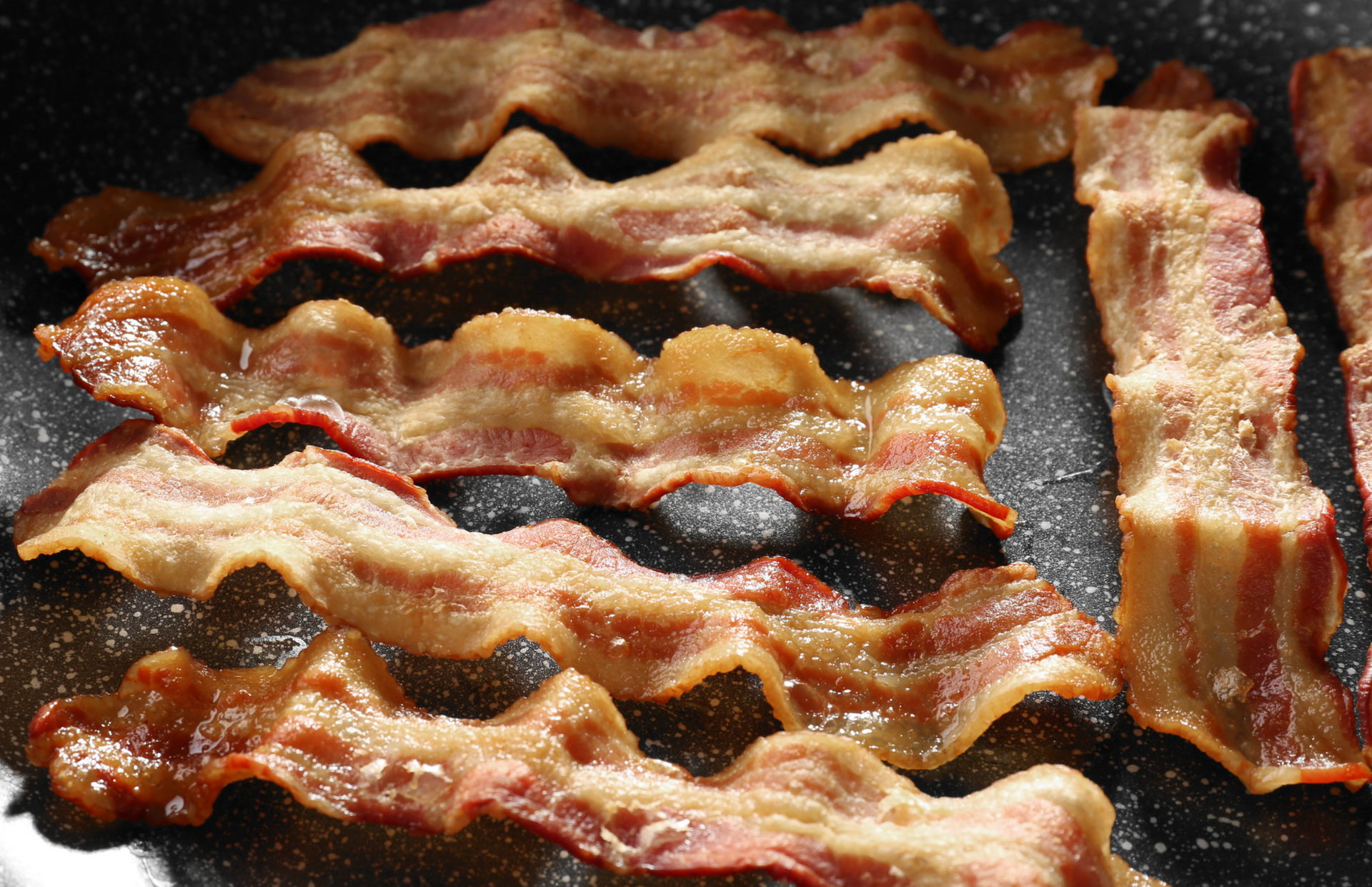 Crispy bacon strips sizzling in a black pan, with golden-brown edges and red meat.
