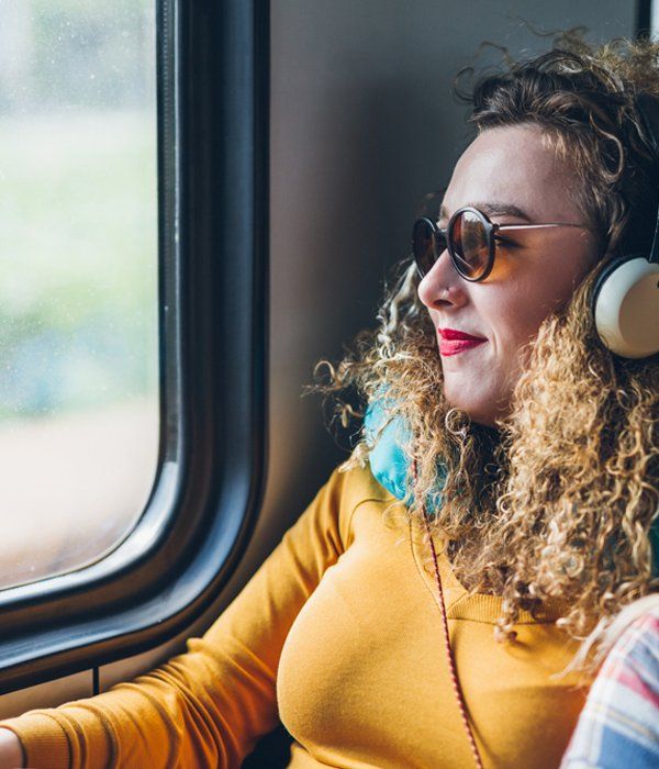 Happy Passenger — Lincoln, NE — OMALiNK Airport Shuttle Charter & Courier Service A person with curly hair, sunglasses, and headphones, wearing a yellow shirt and neck pillow, looking out a train window.