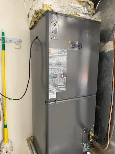 Gray HVAC furnace in a narrow utility closet with exposed pipes and a broom on the wall.
