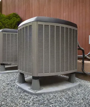 Two outdoor HVAC units on gravel beside a residential fence