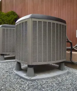 Two outdoor HVAC units on gravel beside a wooden fence and wall