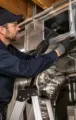 Technician in navy workwear adjusts shiny HVAC ducting in a basement crawlspace while standing on a ladder