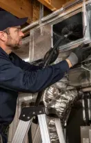 Technician in navy workwear adjusts shiny HVAC ducting in a basement crawlspace while standing on a ladder
