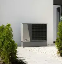Outdoor HVAC unit beside a white house, surrounded by gravel and green shrubs.