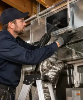 Technician inspecting ductwork in an attic, standing on a ladder and working with gloved hands