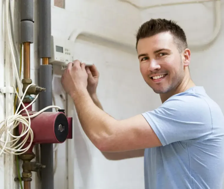 Smiling man adjusting a wall-mounted boiler with exposed pipes and wires