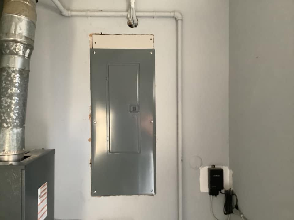 Gray electrical panel on a white wall in a utility room, with ductwork and an outlet nearby