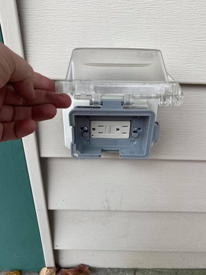 Hand holding open a clear plastic cover over an outdoor electrical outlet on a wall