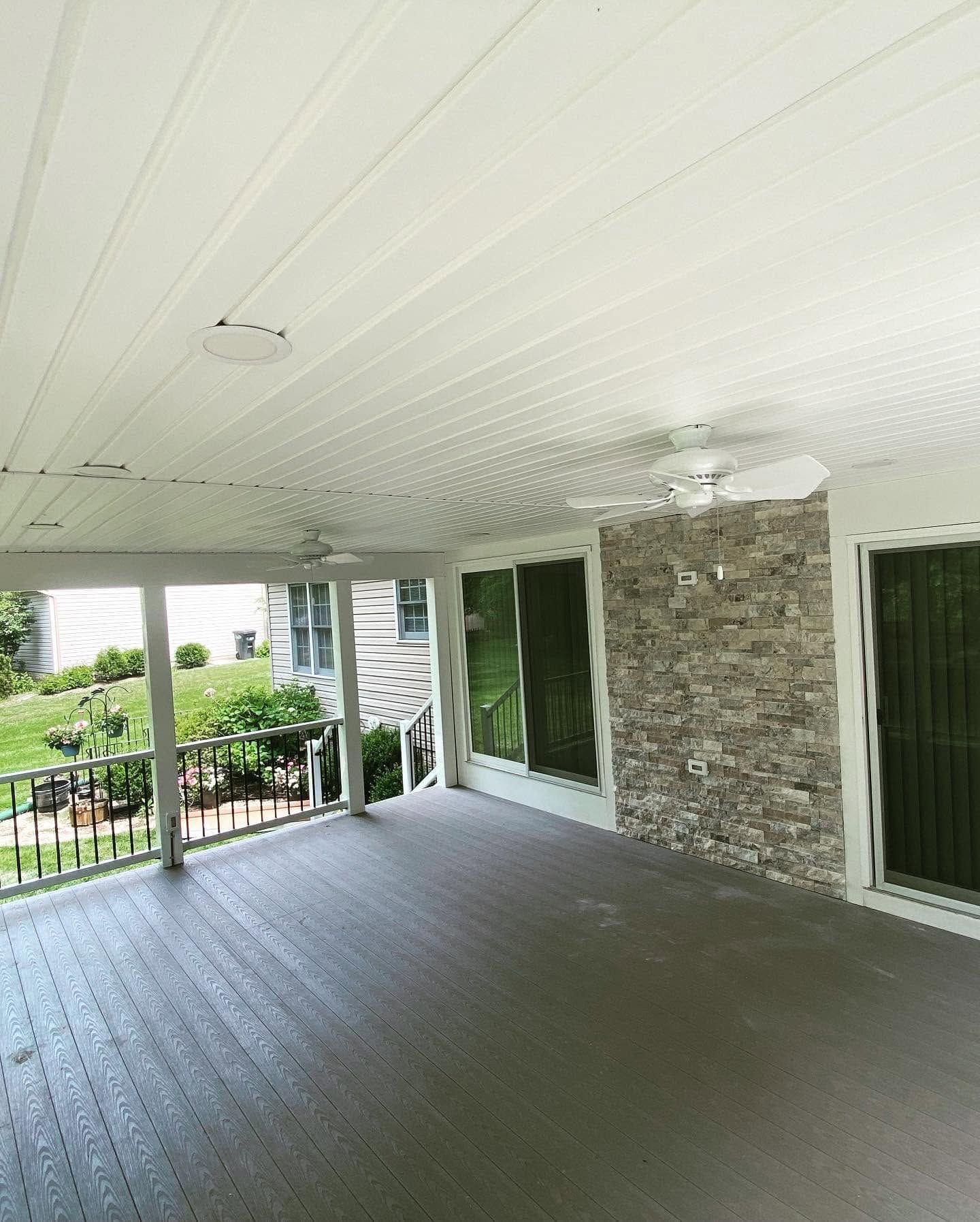 Covered porch with gray flooring, white beadboard ceiling, stone wall, and green outdoor view.