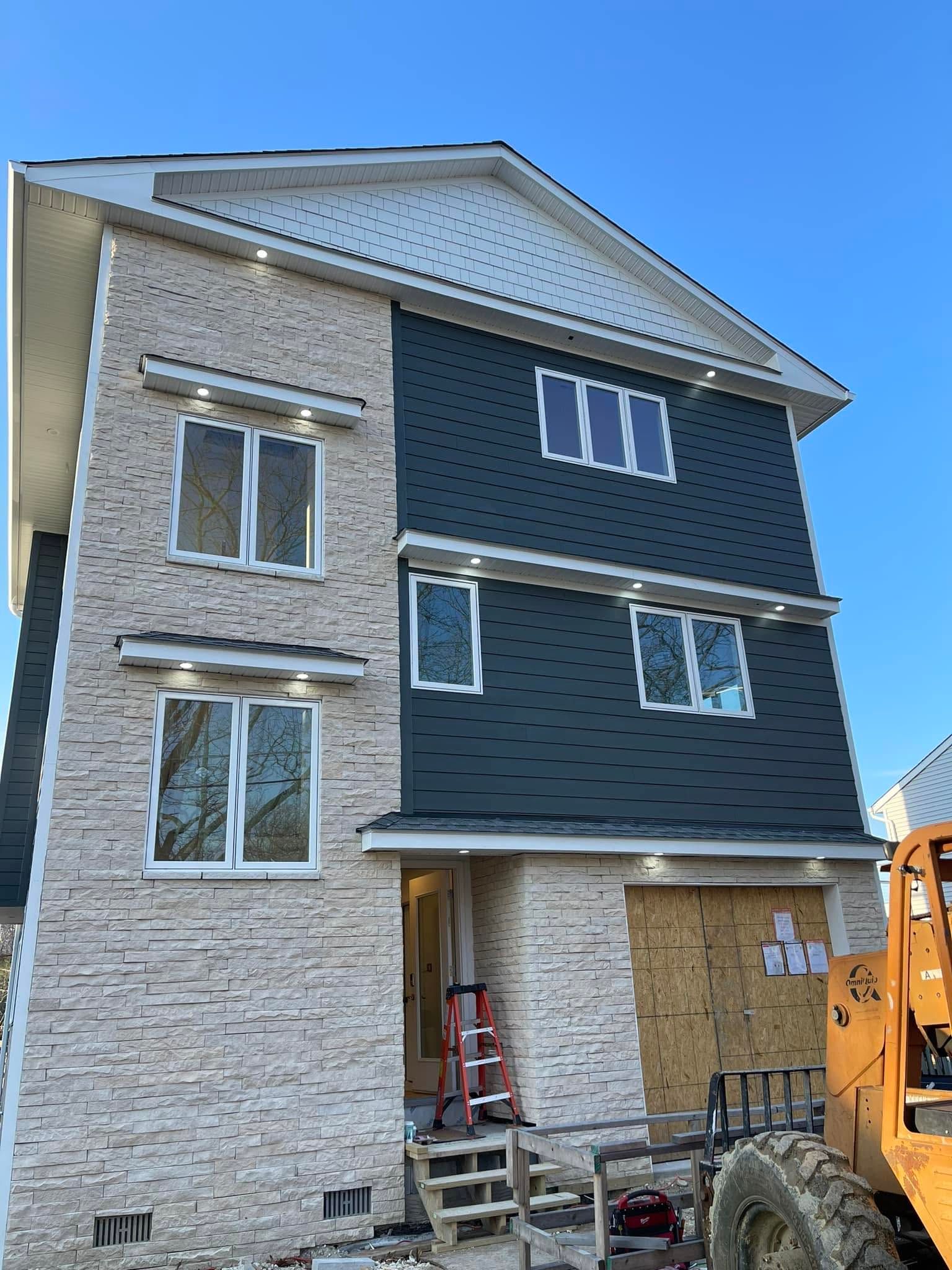 Three-story house under construction with gray siding, stone exterior, and open ground-level entry.