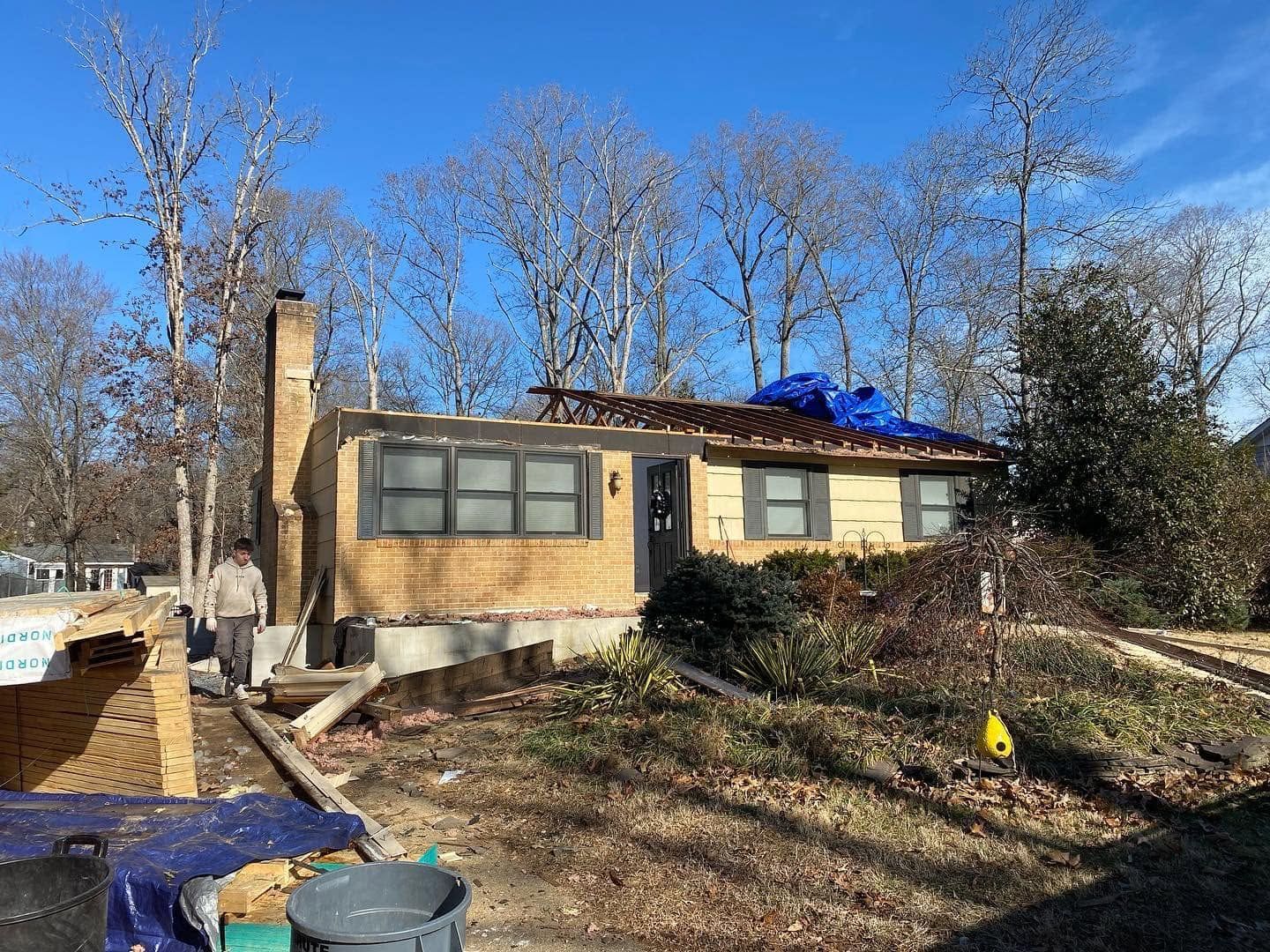 Single-story brick house with boarded windows, damaged roof tarp, and leaf-strewn yard under a blue sky