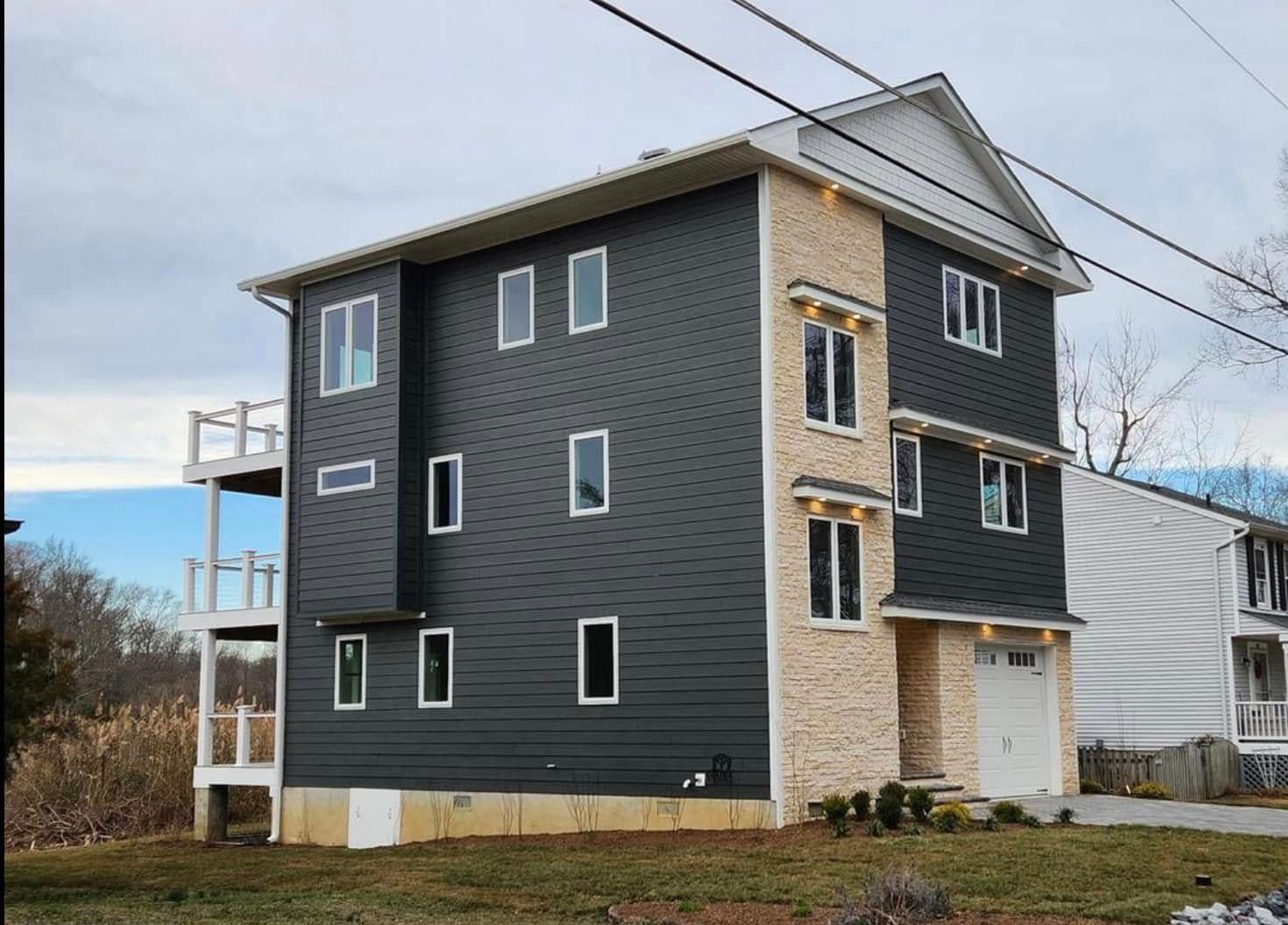 Three-story dark gray house with beige trim and multiple windows under a cloudy sky