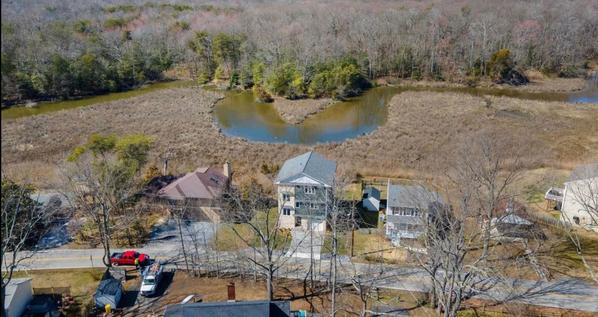 Aerial view of a house by a small pond, surrounded by leafless trees and a wooded area