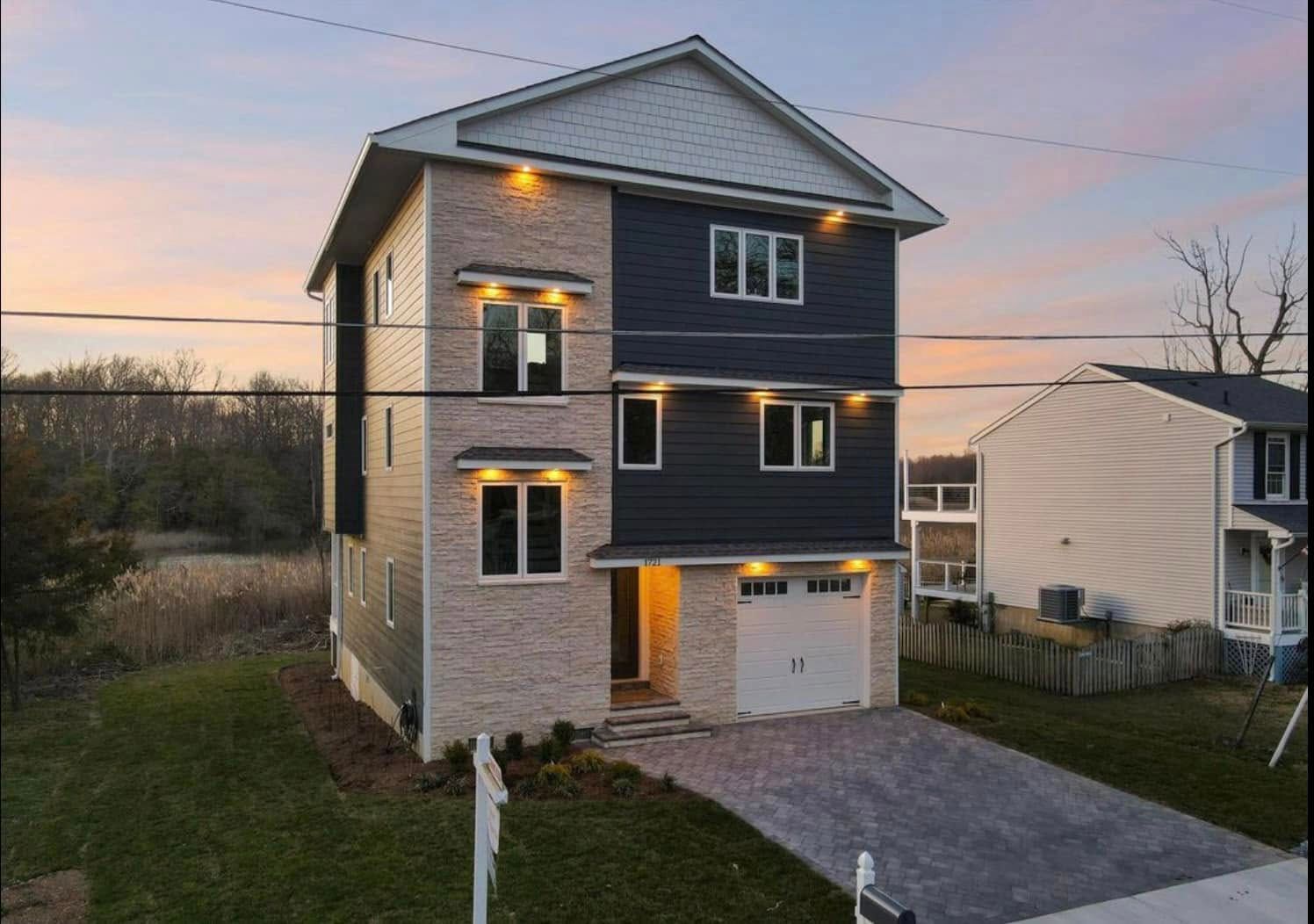 Three-story gray house with a garage at dusk, lit porch lights and a driveway in front.