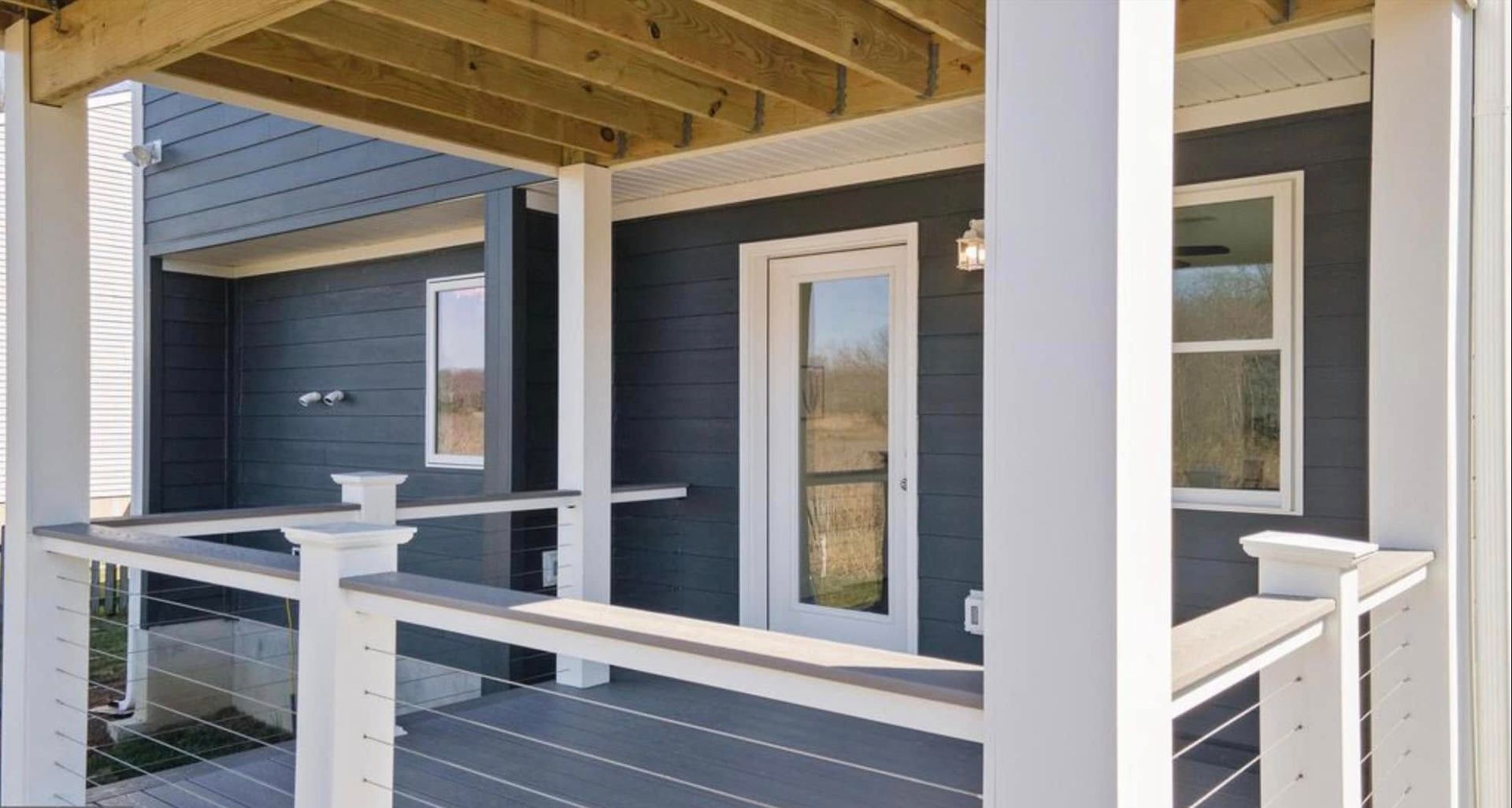 Covered porch with white railings, gray siding, and a glass door under exposed wooden beams.
