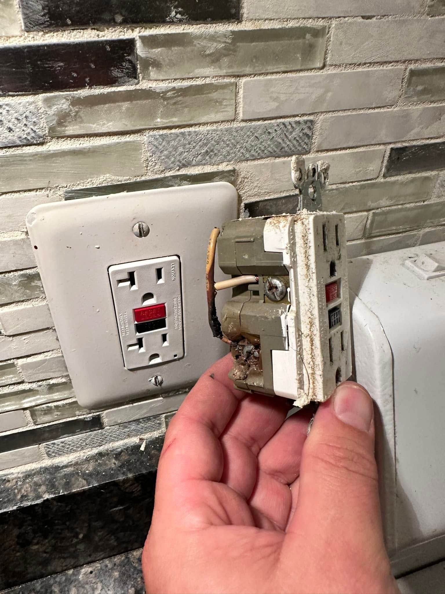 Hand holding a damaged white electrical wall switch beside an outlet and tiled wall