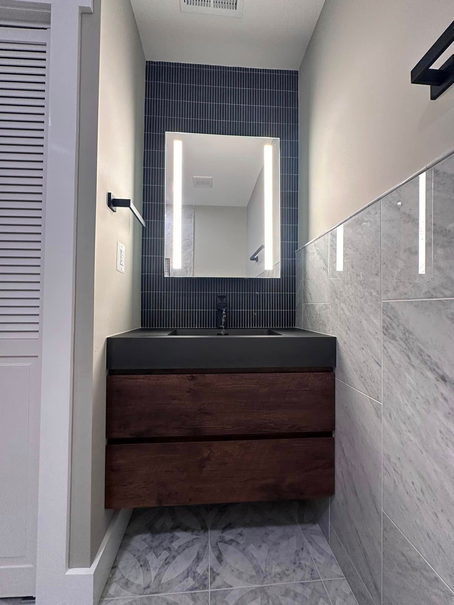 Narrow bathroom vanity with mirror, black sink, wood drawers, and gray tile walls.