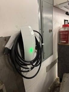 White wall-mounted EV charger with green light and coiled cable beside a gray electrical panel in a garage.
