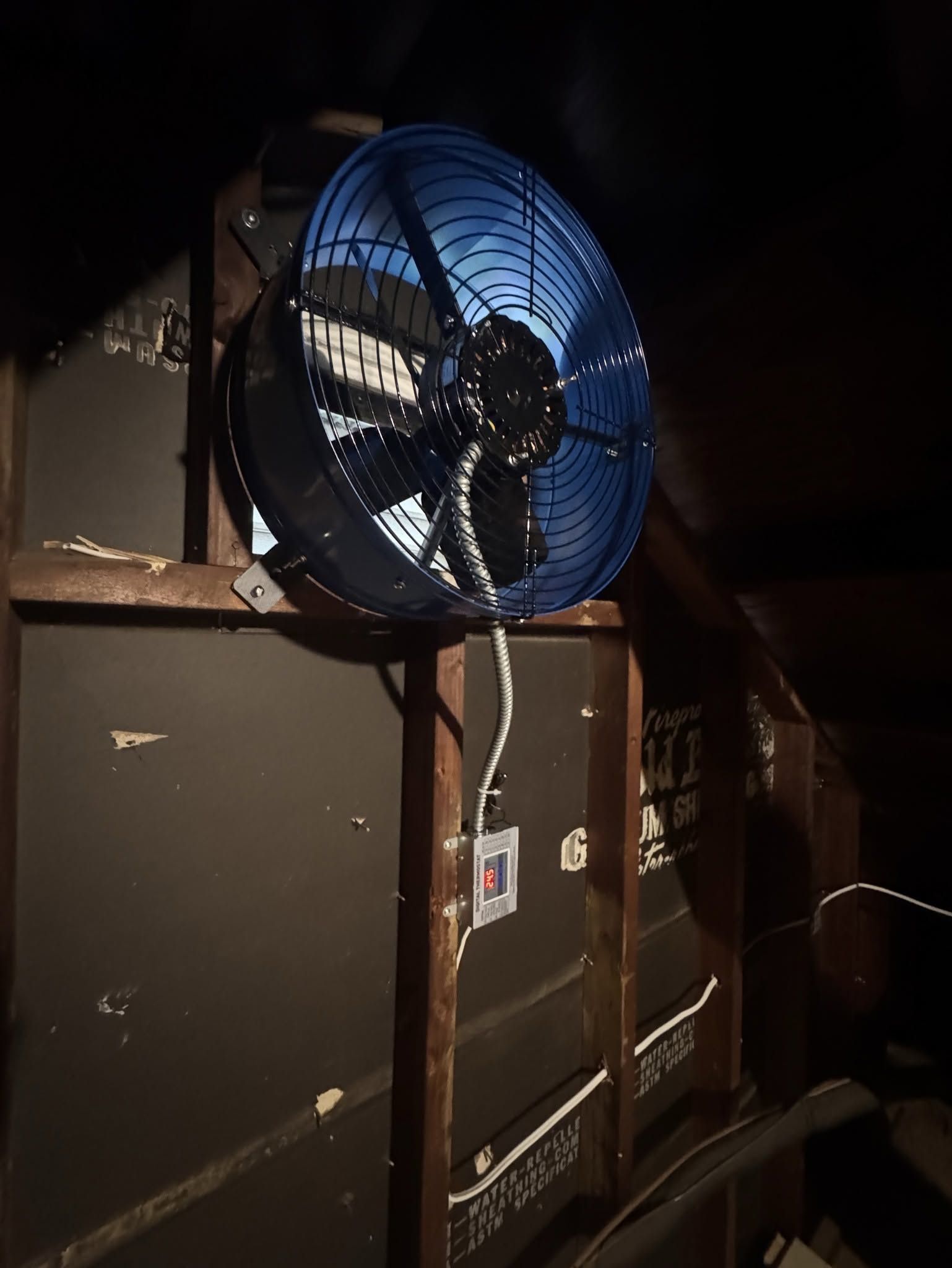 Blue industrial fan mounted on a dark wall, with ducting and cables in a dim utility space