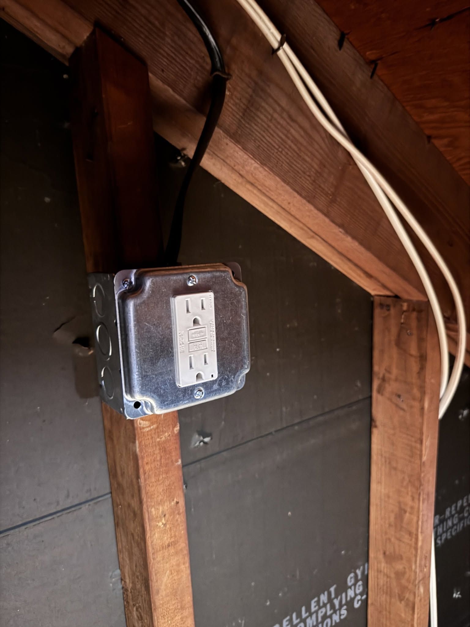 Electrical outlet box mounted on a wooden beam in an attic with exposed wiring.