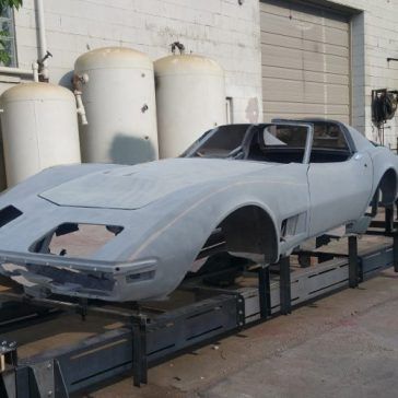 A grey Corvette body on a steel frame, in front of industrial tanks, likely undergoing restoration.