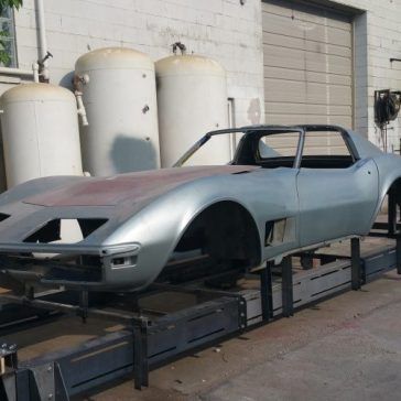 Silver Corvette body on a custom frame, exterior view near tanks and a building.