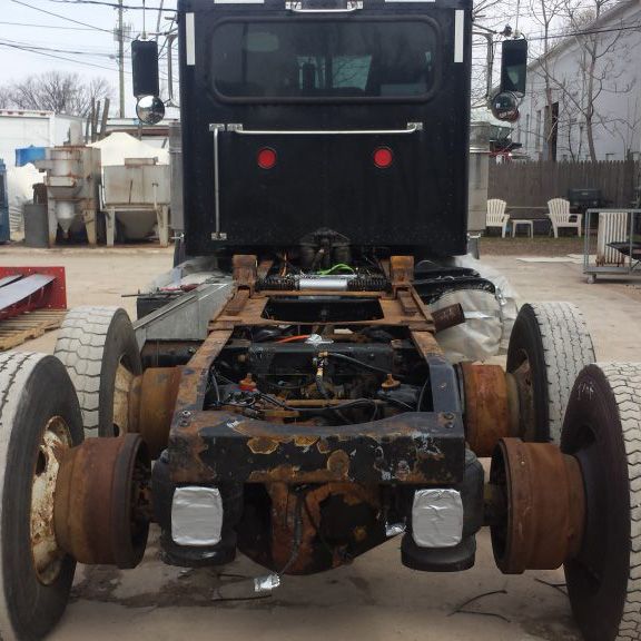 Rear view of a black semi-truck chassis with wheels, rusted frame, and cab. Outdoor setting.