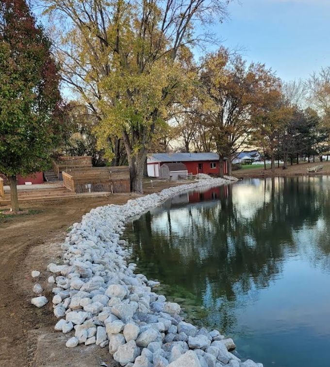 Rock-lined pond shore with calm water, red shed, and autumn trees reflected on the surface