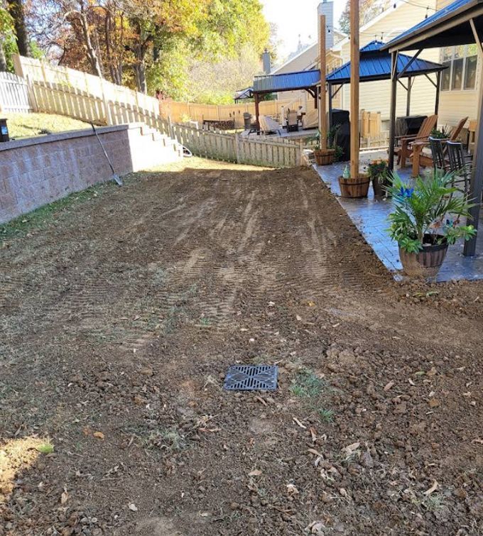 Freshly graded dirt yard beside a house with a patio, fence, and potted plants in daylight