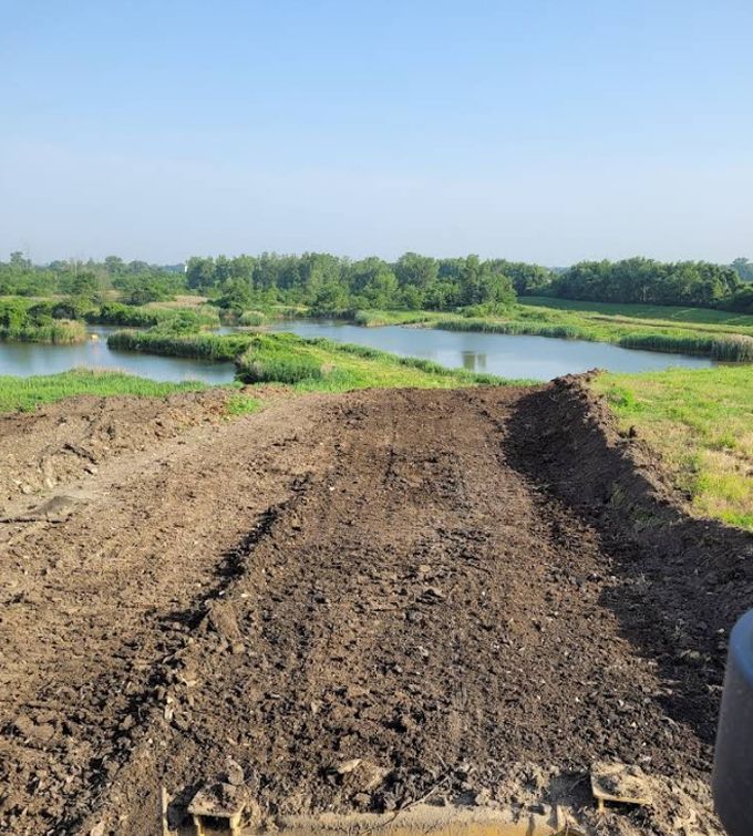 Dirt path leading to a calm river, with green trees and grassy banks under a clear blue sky.