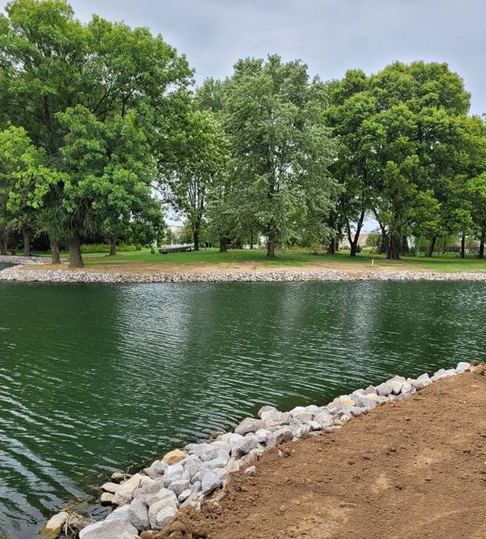 Tree-lined pond with green water, a rocky shore, and a dirt path in the foreground