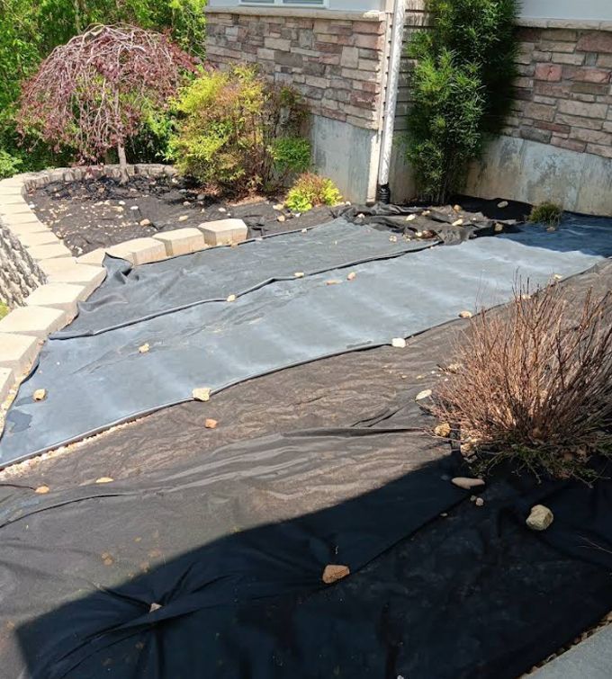 Garden bed covered with black and gray landscape fabric beside a stone house wall and shrubs