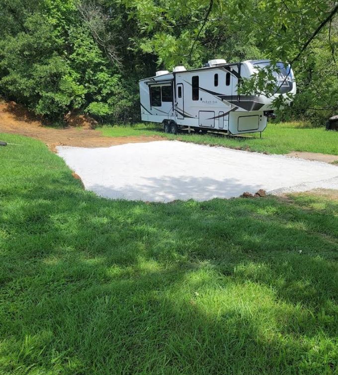 White RV parked beside a paved pad in a grassy, wooded campsite