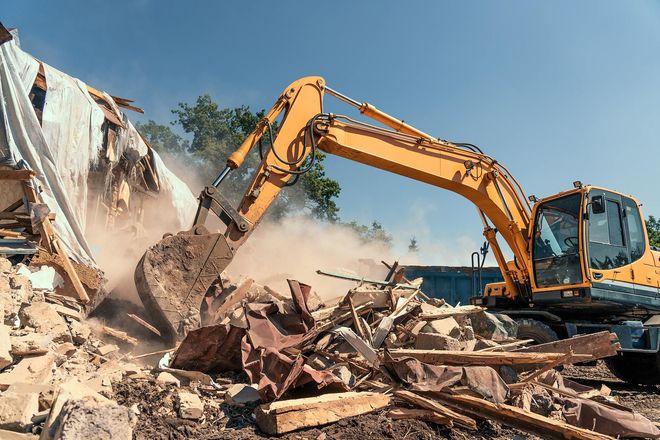 Yellow excavator demolishing a collapsed building, with dust and rubble scattering under a blue sky