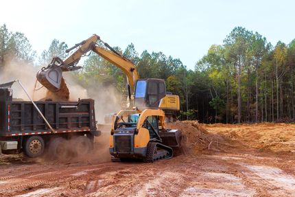 Excavator loading dirt into a dump truck at a muddy construction site with trees in the background