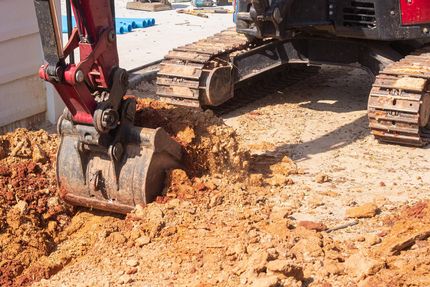 Excavator bucket digging red soil on a construction site with tracked machine in the background