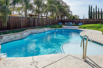 Backyard swimming pool with clear blue water, stone deck, and wooden fence under trees