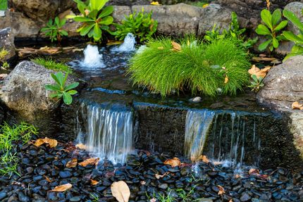 Small garden waterfall flowing over rocks with green plants and pebbles