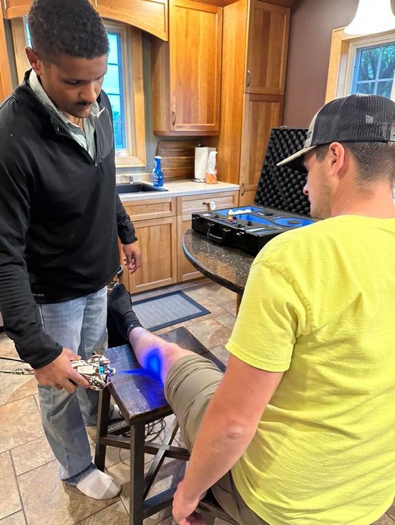 Two people at a kitchen table, one using a blue light device on the other's leg.