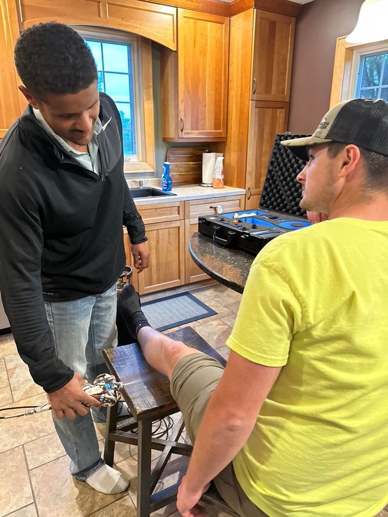 Two people in a kitchen, one seated in a chair while another adjusts his leg.