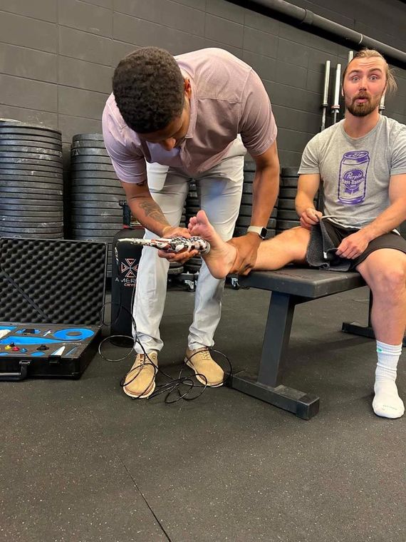 Two men in a gym, one crouching and checking a device while the other sits on a bench.