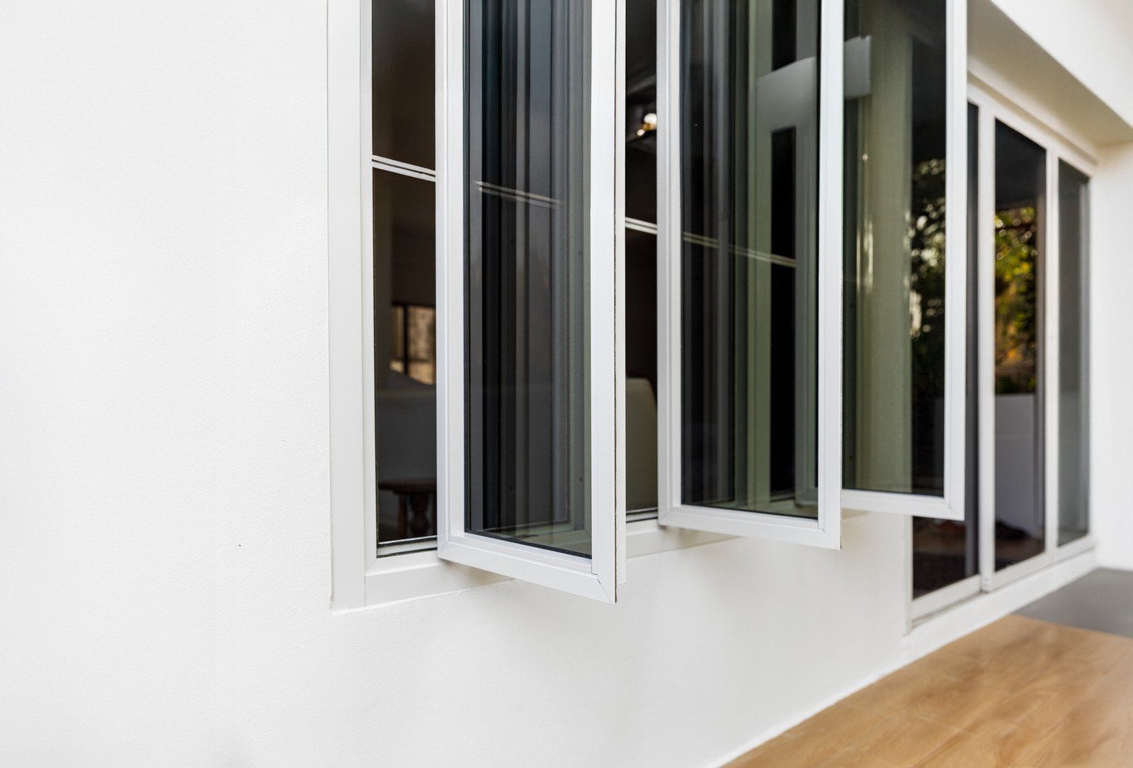 White framed windows with dark screens open outward against a white wall above a wooden floor.