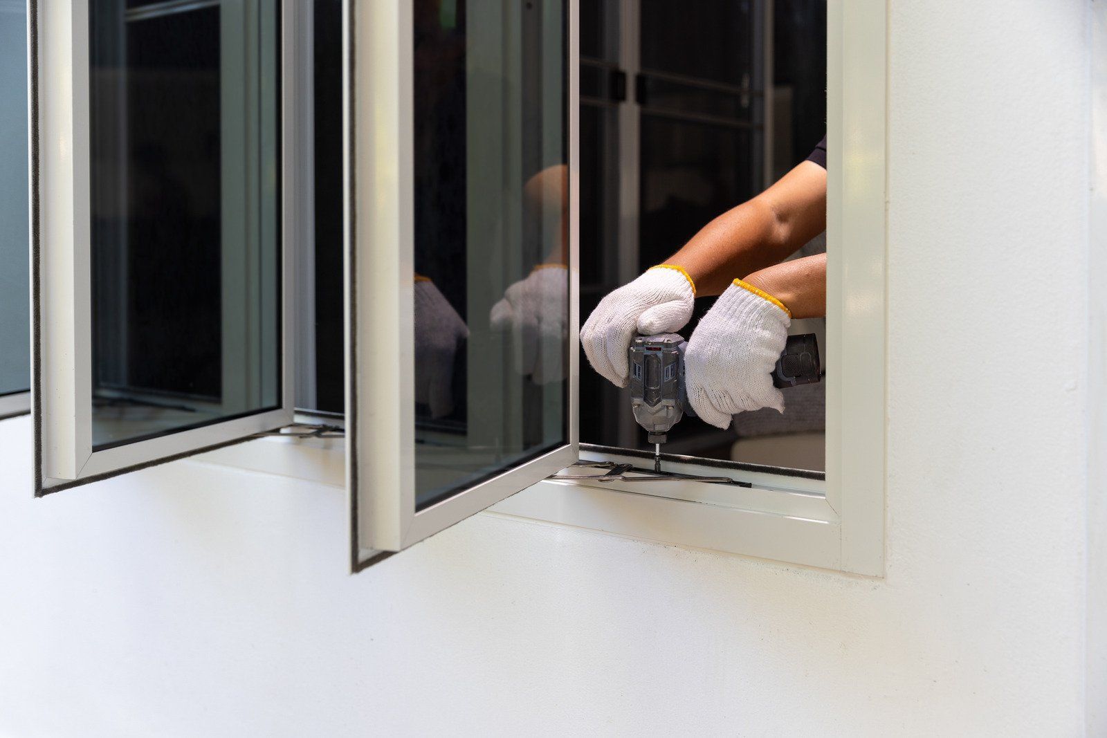 A pair of gloved hands uses a power drill to install or repair a white-framed aluminum window.